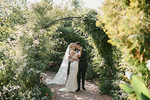 Couple portrait of bride and groom embracing beneath a vine-covered garden arch, her long veil trailing over the brick pathway