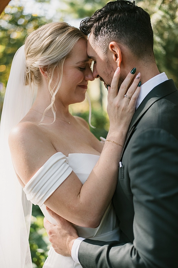 Couple portrait with bride holding groom’s face in a forehead touch wedding photo, veil flowing in sunlit greenery with bokeh trees