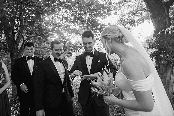 Wedding ceremony moment as groom places ring on bride’s finger, bride in veil holding bouquet under leafy trees in dappled sunlight