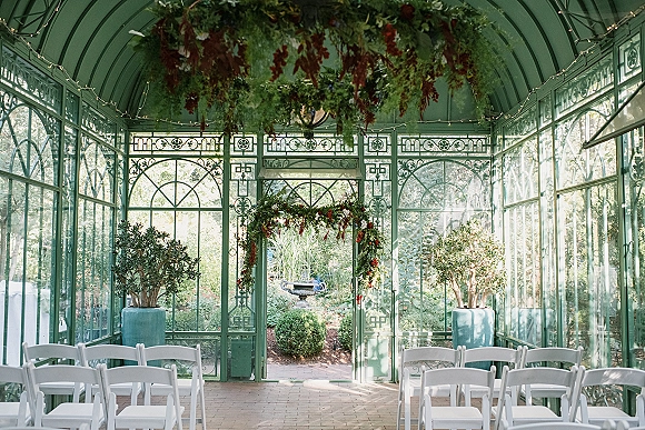 Ceremony setup with greenhouse wedding ceremony seating, white folding chairs lining a brick aisle toward a floral arch in a glass conservatory