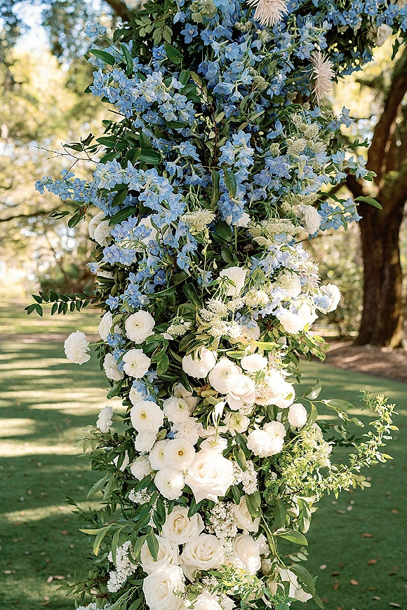 Wedding floral installation ceremony floral pillar with white roses, ranunculus, and blue delphinium in a sunlit garden lawn setting