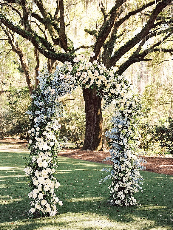 Wedding ceremony arch with white roses, blue flowers, and greenery in a sunlit garden lawn beneath large trees