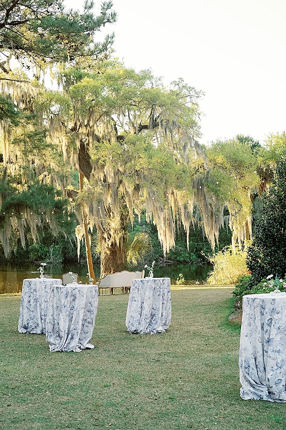 Outdoor reception setup with wedding cocktail hour setup, blue floral cocktail tables, lounge seating, and string lights beneath mossy trees by water