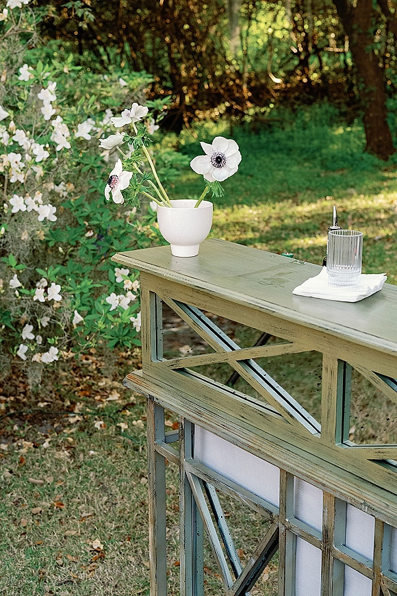 Wedding bar setup with a white vase of flowers, glass tumbler, napkin, and bottle pour spout on a bar counter in a garden setting