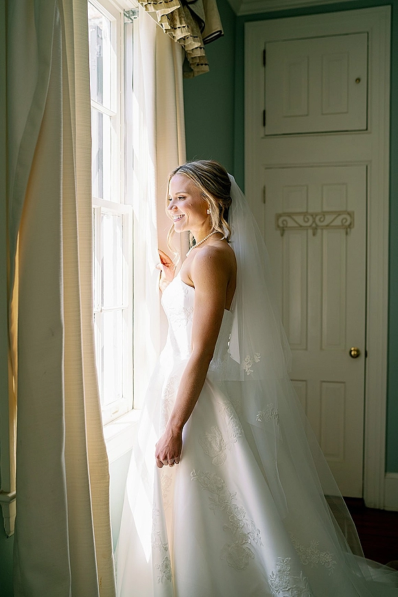 Bridal portrait of a bride by window in natural light, holding the curtain in side profile with a long veil and pearl necklace indoors