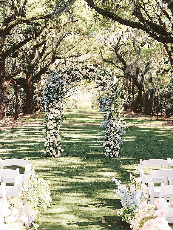 Ceremony setup with outdoor wedding ceremony floral arch of white roses, blue flowers and greenery along an oak-lined aisle with moss
