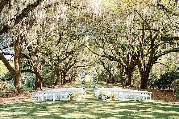 Ceremony setup with outdoor ceremony seating of white folding chairs facing a floral arch along a tree-lined aisle under oak trees with Spanish moss