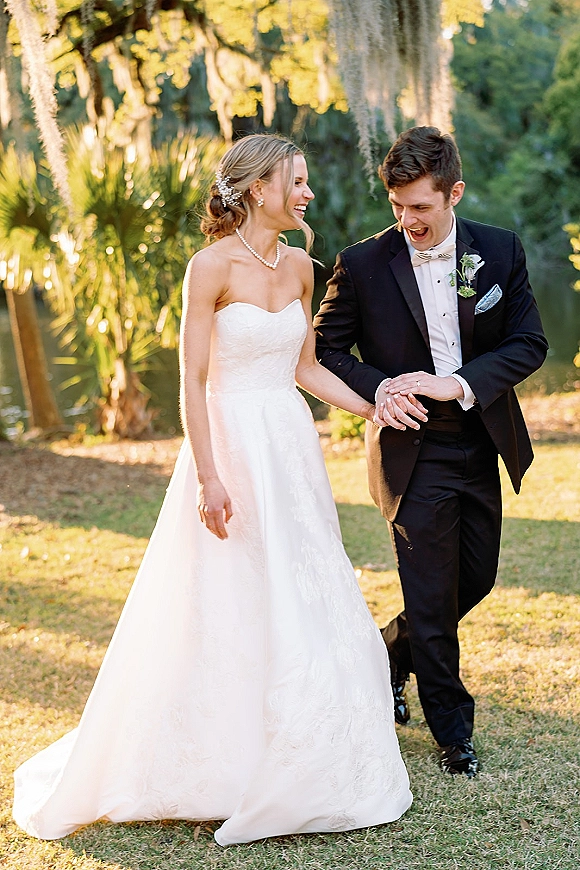Couple portrait of bride and groom laughing hand in hand on a garden lawn under hanging moss in golden hour light, pearl necklace accent