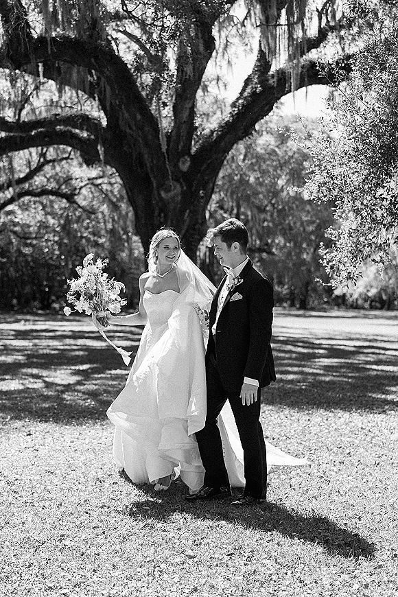 Couple portrait of bride in strapless wedding dress with veil and bouquet beside groom in tuxedo under sunlit oak with hanging moss