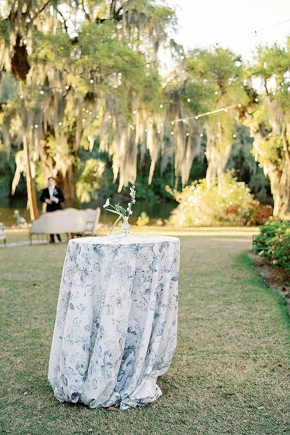 Cocktail table decor with a blue floral tablecloth, bud vase of white flowers and greenery, under string lights on a lakeside lawn