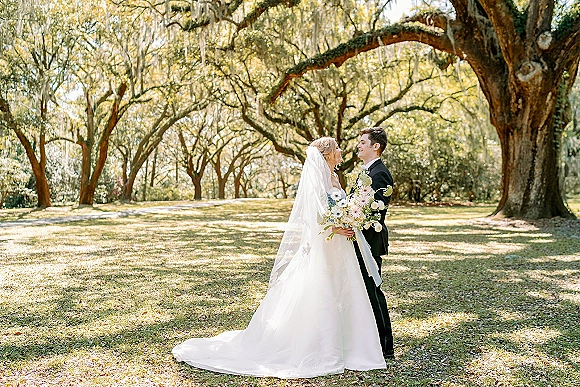 Couple portrait of bride and groom face to face, bride holding bouquet and veil flowing beneath oak trees with hanging moss in sunlit lawn