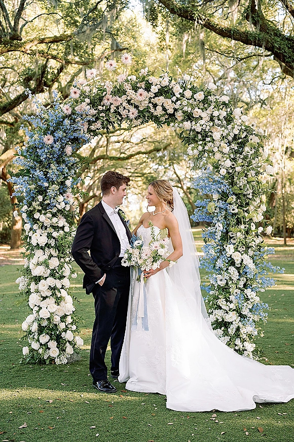 Couple portrait of bride and groom under floral arch, smiling in sunlight, with blue-white bouquet, veil and tuxedo beneath oak trees with moss