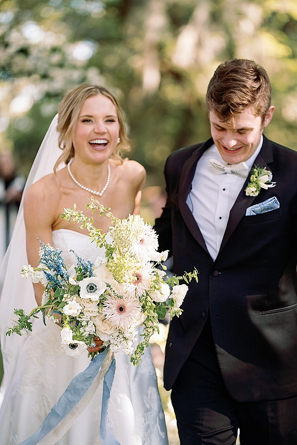 Wedding couple portrait of bride and groom walking in a garden, bride laughing in veil with blue bouquet, groom in tux looking down