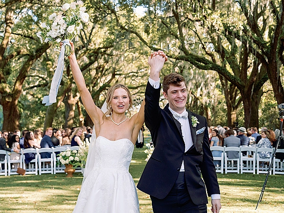 Recessional moment as newlyweds walk down the aisle holding hands, bride lifting bouquet overhead beside groom in tuxedo under oak trees