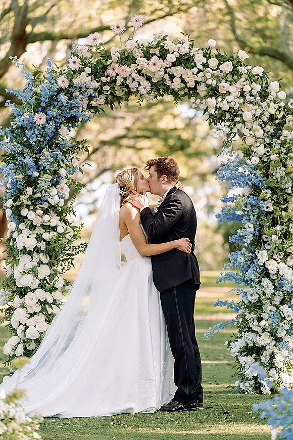 Wedding kiss as the couple kissing under arch of white roses, blue flowers, and greenery, sunlight on a garden lawn with trees