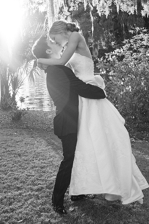 Wedding kiss as the groom lifts the bride in a dip kiss wedding photo, her strapless dress and updo framed by lakeside mossy trees
