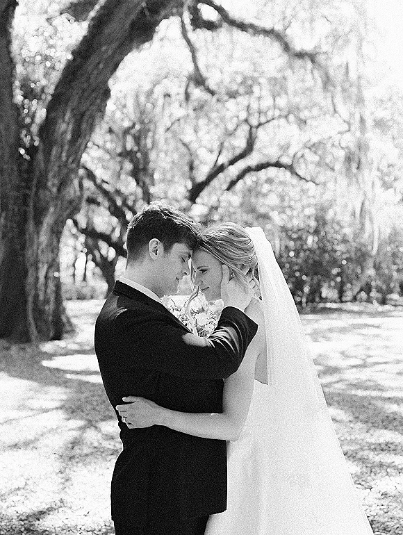 Wedding couple portrait in black and white with bride and groom embracing, foreheads touching under trees in dappled park light, veil flowing