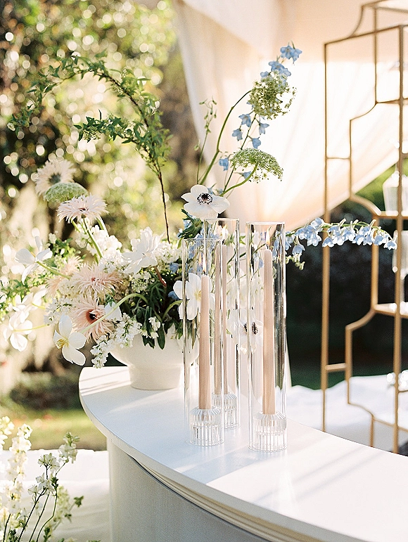 Ceremony altar decor with wedding ceremony backdrop of white and blue florals, taper candles in glass vases, and a gold geometric frame in a garden setting
