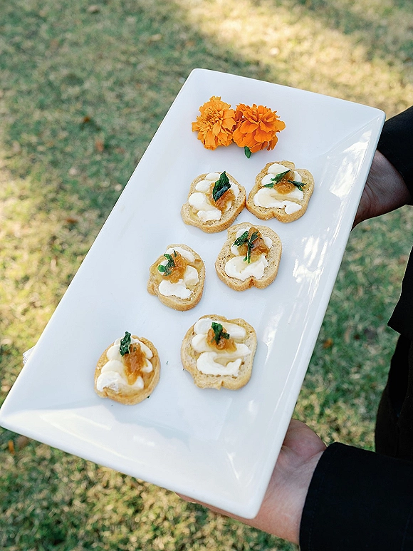 Wedding appetizers on a white platter with crostini wedding appetizers topped with creamy cheese, chutney, herbs, and orange flowers outdoors on grass lawn