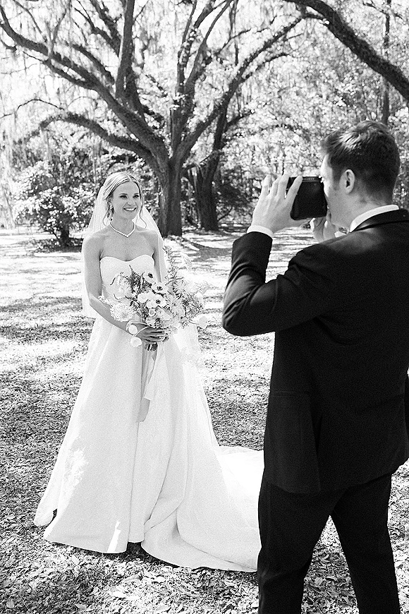 Bridal portrait in a black and white wedding photo, bride in strapless gown and veil holding a bouquet as groom photographs her in a park with tall trees