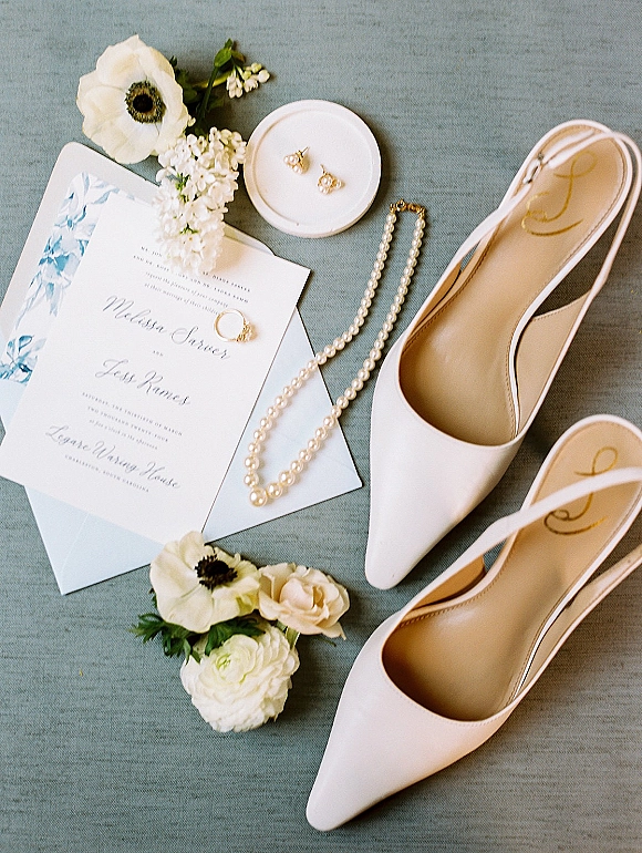 Wedding invitation suite with blue and white invitations styled alongside rings, pearls, heels, and flowers on linen fabric flat lay