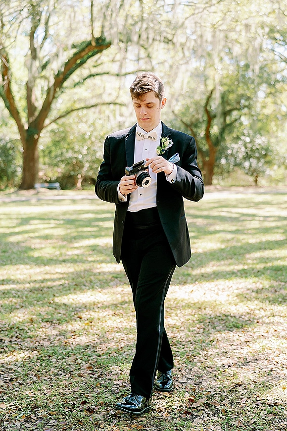 Groom portrait with groom holding camera, wearing a black tuxedo and boutonniere on a grassy lawn beneath oak trees with Spanish moss