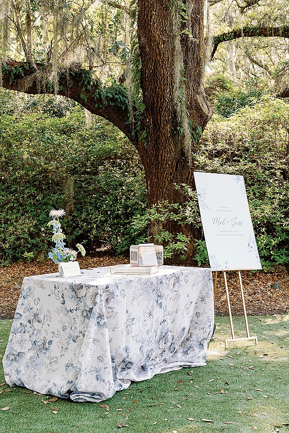 Wedding welcome table with a wedding welcome sign on an easel, guest book and card box on a blue floral tablecloth beneath trees