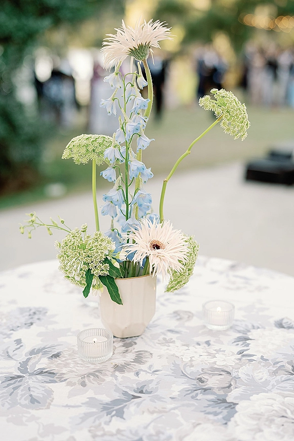 Wedding centerpiece with blue-and-white wildflowers in a white vase on a patterned tablecloth, with votive candles on an outdoor lawn