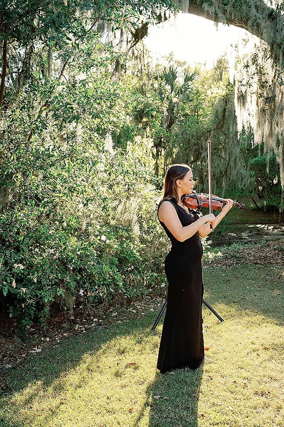 Wedding ceremony musician, wedding violinist playing violin beside a music stand on a sunlit garden lawn under trees with spanish moss
