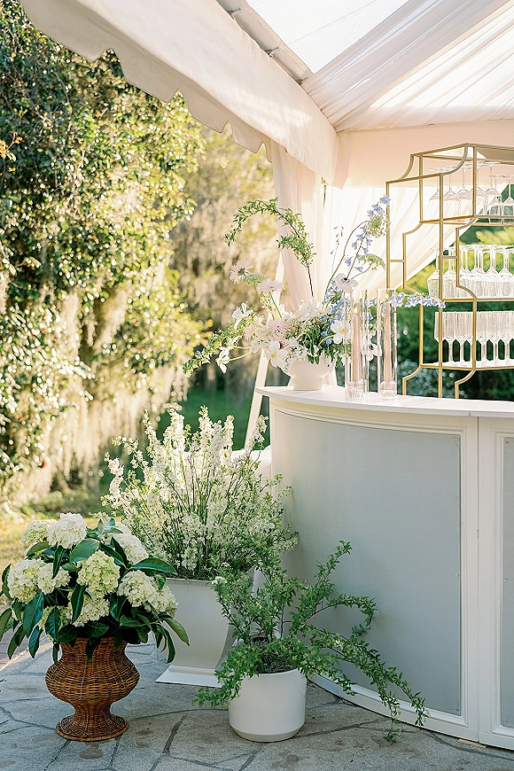 Wedding bar setup with white mobile bar, gold shelving, hanging wine glasses, and floral greenery under a canopy on a sunny stone patio