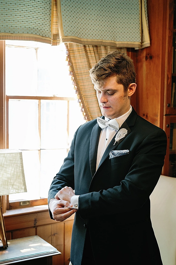 Groom portrait in a black tuxedo adjusting cufflinks with a white boutonniere in soft window light beside plaid curtains and wood paneling