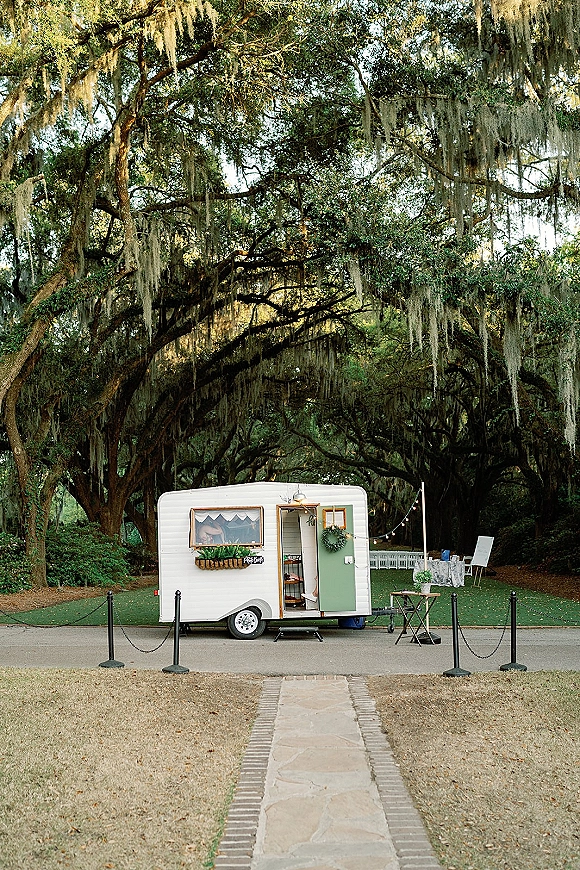 Wedding photo booth vintage camper photo booth with string lights, chalkboard sign, and wreath under mossy oak trees on a lawn path