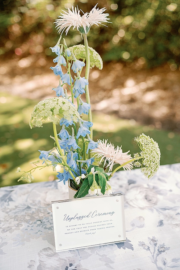 Unplugged ceremony sign in acrylic frame with calligraphy, beside blue delphinium and white mums on a patterned table in a sunny garden