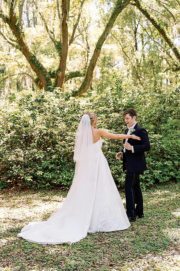 First look moment as bride in a strapless gown and veil reaches for groom in tuxedo, amid dappled sunlight in a garden backdrop