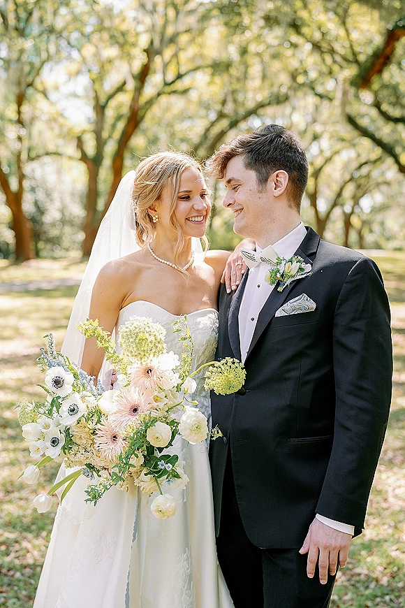 Couple portrait of bride and groom smiling, bride holding bouquet with veil and pearls, groom in black tuxedo beneath oak trees