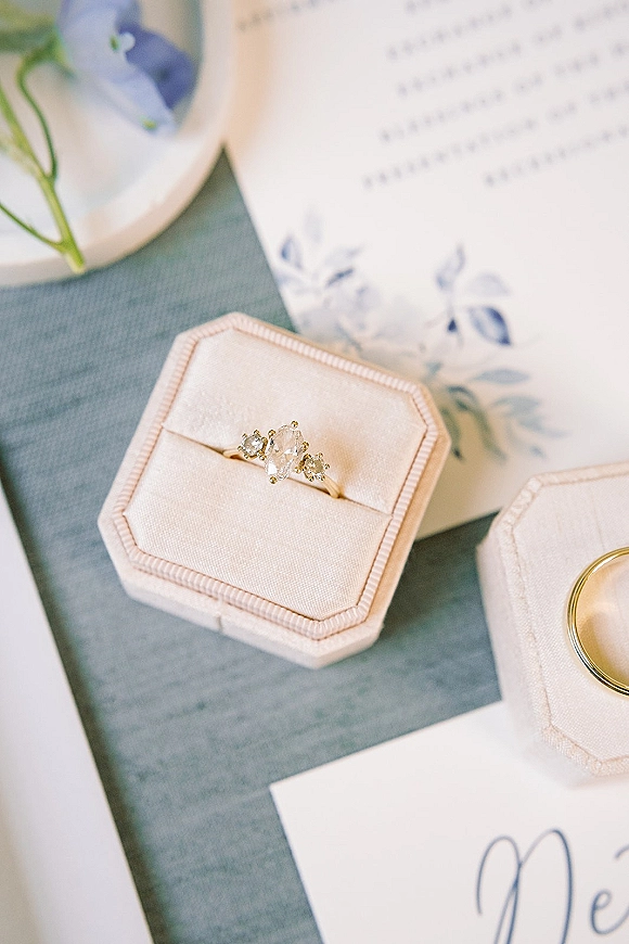 Engagement ring and oval engagement ring atop a calligraphy invitation suite on linen, with velvet ring box and blue floral stationery nearby