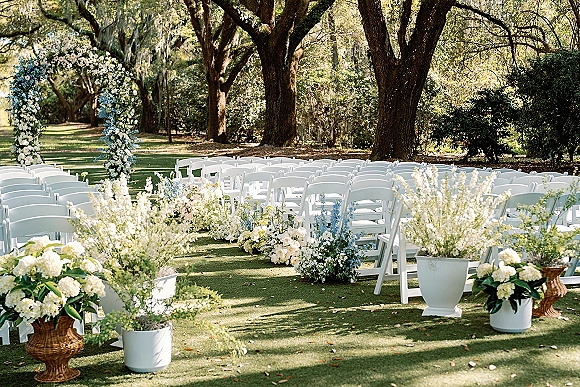 Outdoor ceremony setup with garden wedding ceremony seating rows of white folding chairs facing a floral arch on a lawn under tall trees