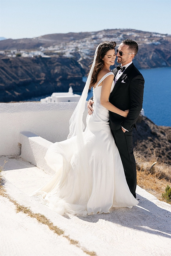 Couple portrait of bride and groom hugging, her veil blowing as he wears sunglasses beside white stucco wall with ocean cliffs backdrop
