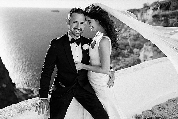 Couple portrait in a black and white wedding portrait, bride leaning on groom as her veil blows by ocean cliffs and stone railing