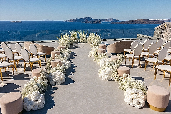 Ceremony aisle decor with outdoor ceremony setup of white hydrangeas and baby’s breath, gold chairs and sofas on a cliffside ocean-view terrace