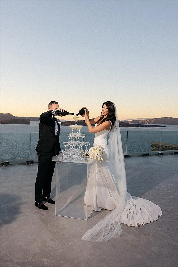 Champagne tower as newlyweds pour wedding champagne tower on a clear acrylic table, with ocean-view terrace and sunset cliffs behind