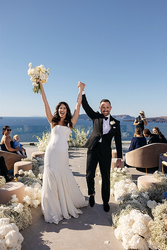 Wedding recessional as bride raises bouquet and groom in tux walks aisle with hydrangeas and petals on an ocean-view cliffside terrace