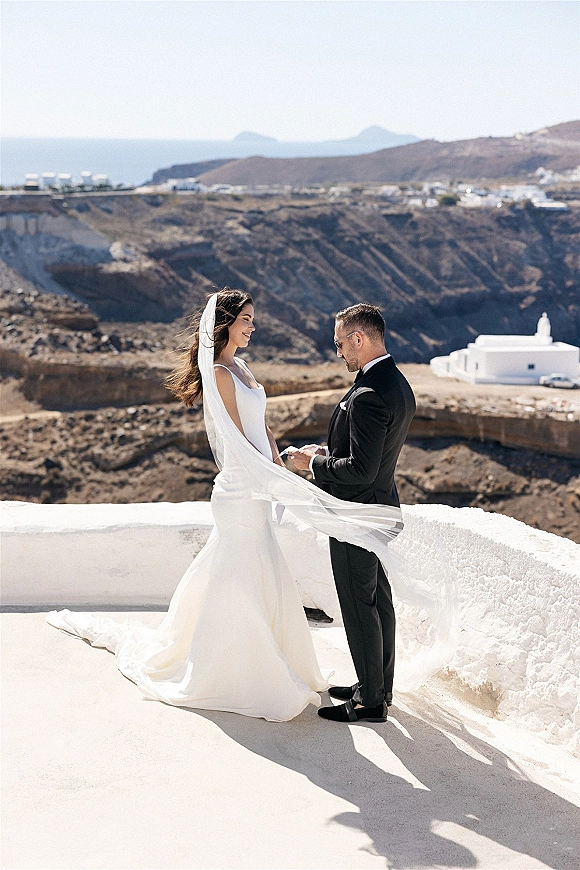 Couple portrait of bride and groom holding hands, her veil blowing in wind as they stand by a cliffside ocean view in sunglasses
