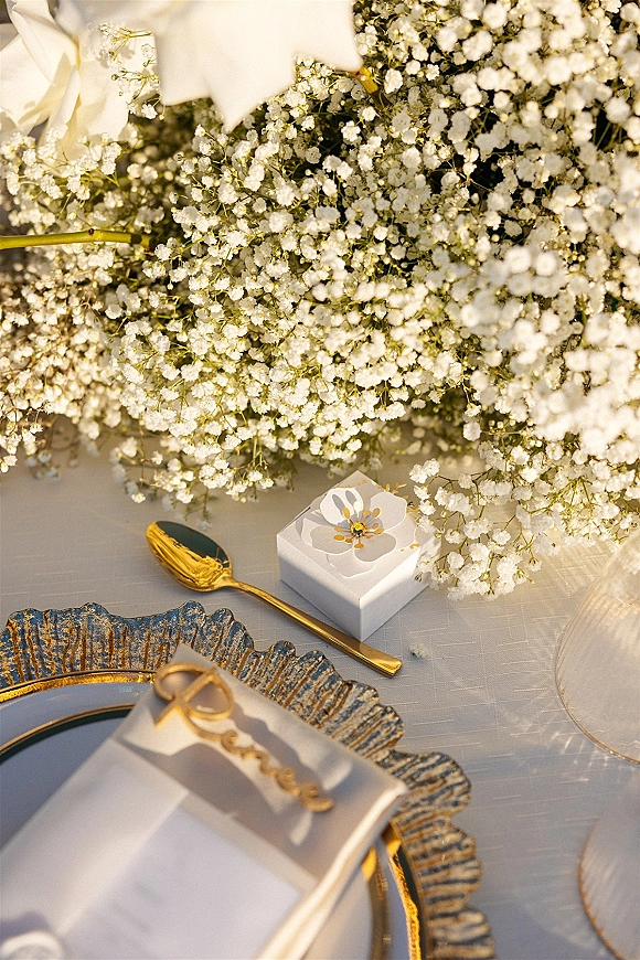 Wedding place setting with gold flatware, scalloped charger plate, baby's breath accent, menu card and favor box on a white tablecloth in sunlit shadows