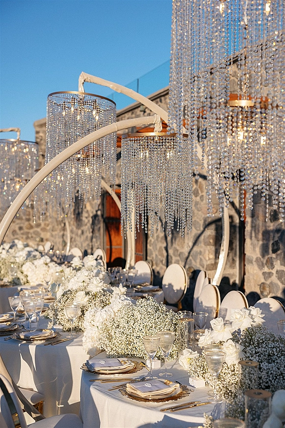 Reception tablescape on an outdoor reception table with white roses and baby's breath beneath crystal chandeliers and bead curtains on a terrace