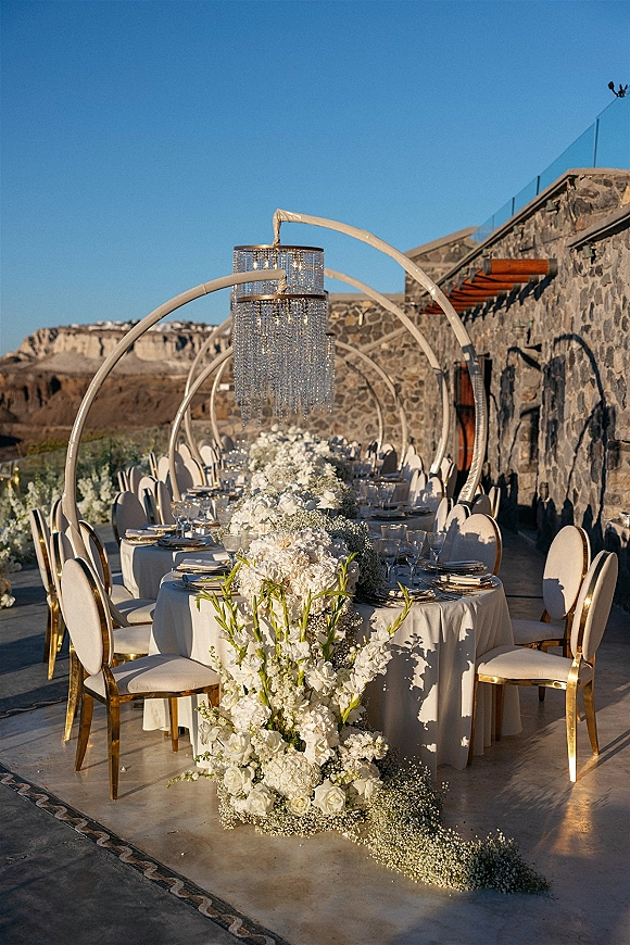 Reception tablescape at an outdoor wedding reception with round tables, white floral centerpieces, crystal chandelier, and terrace stone wall backdrop
