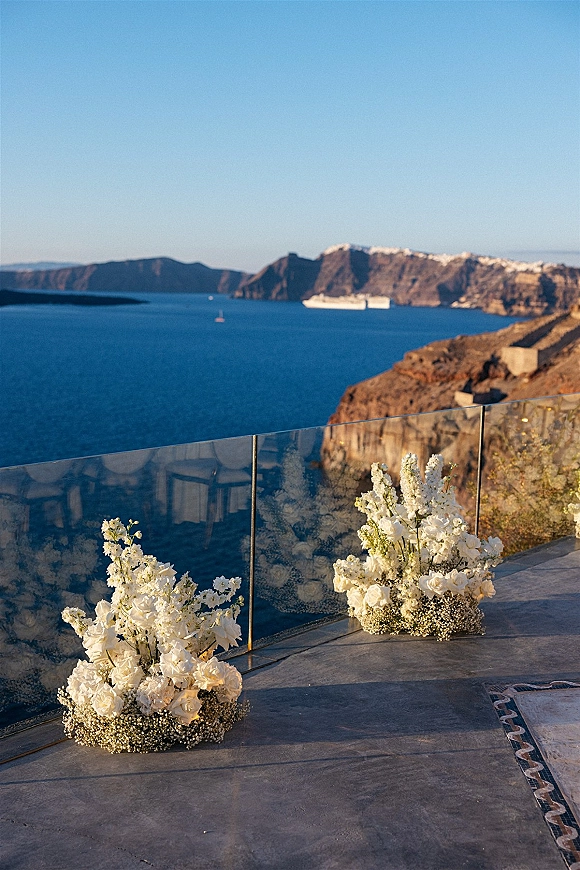 Ceremony aisle decor with low white rose and baby's breath arrangements along a stone patio, set beside a glass railing with ocean cliffs beyond