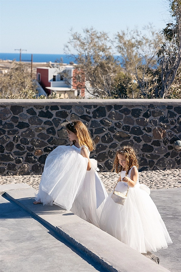 Flower girls in white tulle flower girl dresses walk with a basket along a paved walkway by a stone wall with coastal views and blue sky