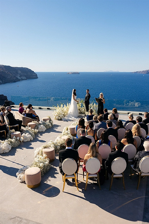 Wedding ceremony with white floral arrangements and aisle flowers as bride and groom exchange vows on a terrace with ocean view and glass railing