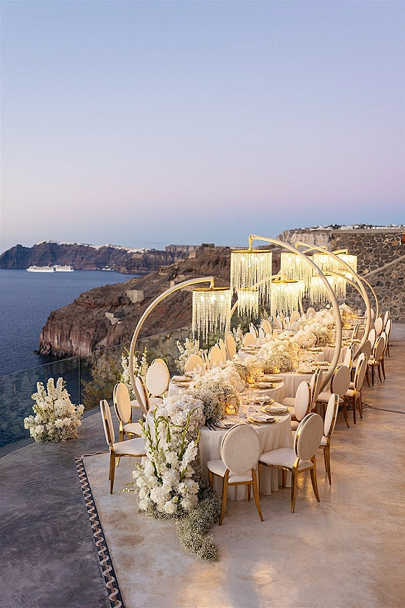 Reception tablescape for an outdoor wedding reception with white floral centerpieces, candles, and gold chargers on a cliffside terrace at sunset